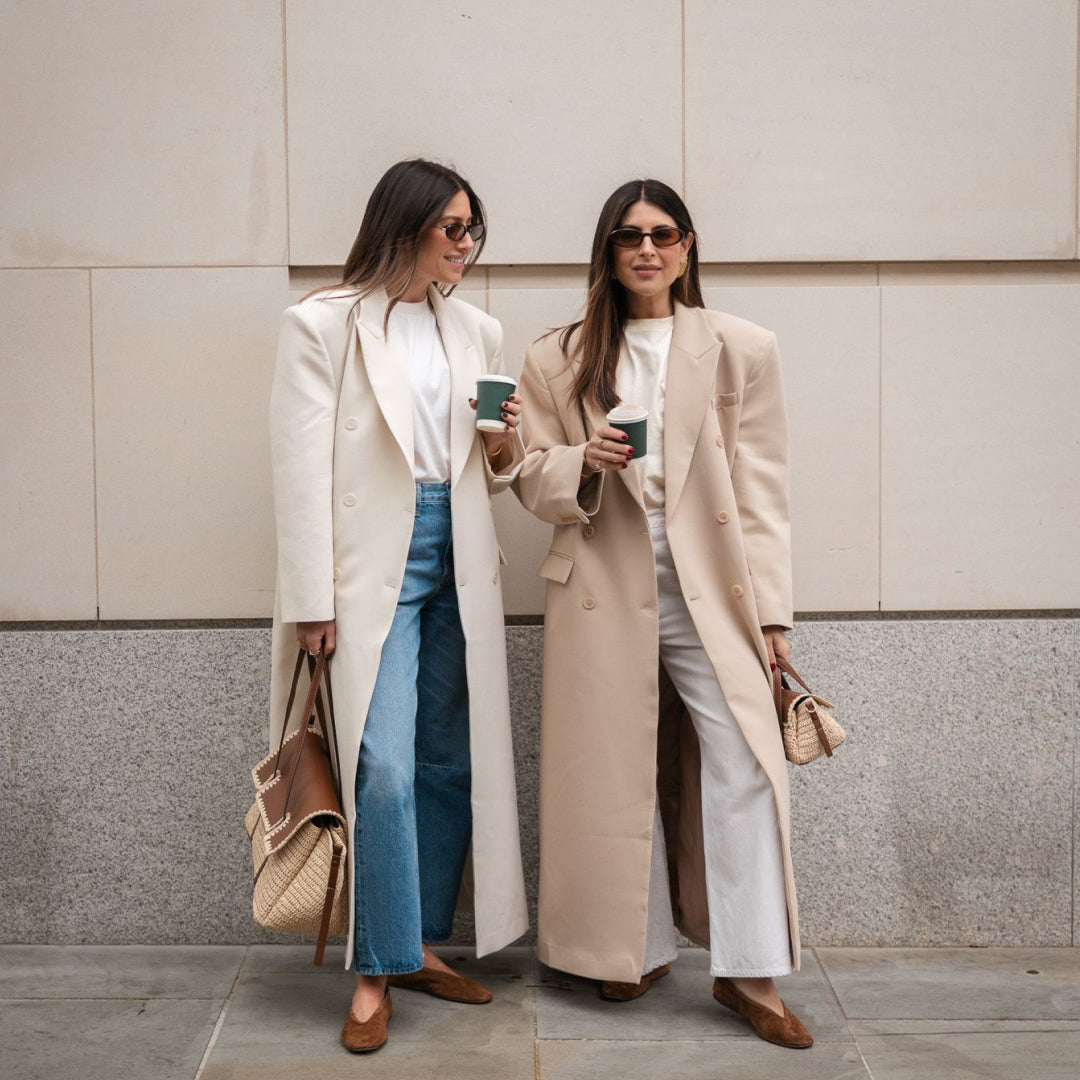 Two women in long coats standing against a neutral wall.