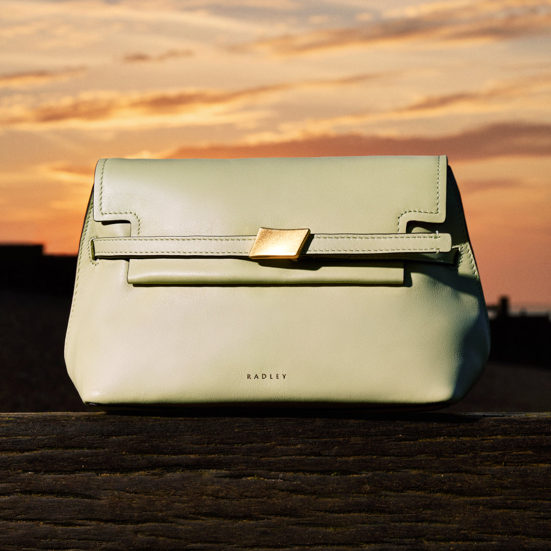 Light green handbag with gold clasp on a wooden surface against a sunset sky